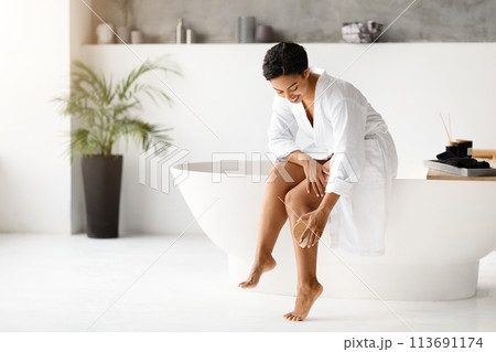 Smiling black woman in bathrobe dry brushing legs, practicing skin exfoliation routine 113691174