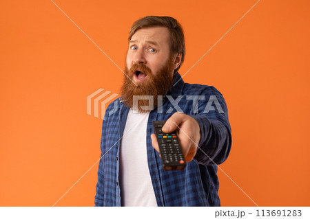 emotional bearded guy holding remote control watching tv, orange backdrop 113691283