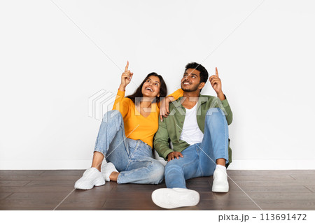 Smiling indian couple pointing up at free space while sitting on floor Smiling indian couple pointing up at free space while sitting on floor 113691472