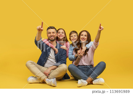 Happy family pointing up sitting together on yellow background Happy family pointing up sitting together on yellow background 113691989