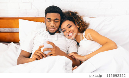 Smiling black millennial couple using smartphone on bed 113692322