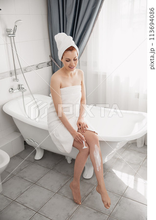 Young woman shaving her legs in bath. Woman with towel over head, resting in bathtub shaving leg. Lady using razor, while sitting in bathtub. Body care concept. Skin body care. Girl in shaving legs 113694086