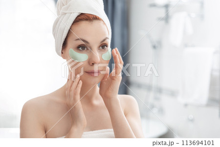 Woman wrapped in towel is examining her face in mirror, focusing on her skin, lips, eyebrows. She smiles at her reflection. Girl with towel on head takes care of her facial skin 113694101