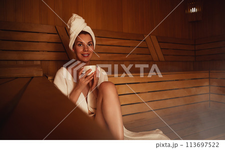 Young woman relaxing and sweating in hot sauna wrapped in towel. Girl In Sauna. Interior of Finnish sauna, classic wooden steam room with hot steam. Russian bathroom. Relax in hot Bathhouse with steam 113697522