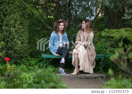 Good-looking girl friends talking and sitting on a park bench outdoors Good-looking girl friends talking and sitting on a park bench outdoors 113701373