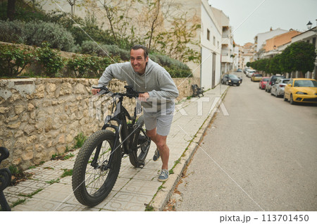 European young man pushing his electric mountain bike up the street. Active lifestyle. Sport. Adventure travel on bicycle. Using el-bicycle ad ecologically friendly mode of urban transport European young man pushing his electric mountain bike up the street. Active lifestyle. Sport. Adventure travel on bicycle. Using el-bicycle ad ecologically friendly mode of urban transport 113701450