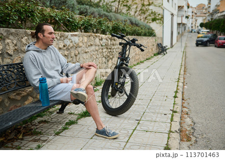 Young man cyclist sitting on a bench near his electric bike, relaxing after ride in mountain city 113701463