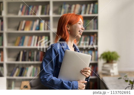 Happy red-headed girl holding laptop standing in college library 113702691