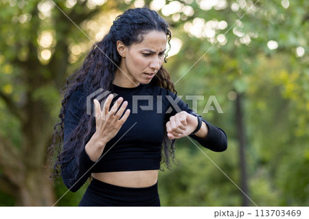 A confused Hispanic woman in workout attire checks her watch during an exercise session in a park, expressing uncertainty and slight frustration. 113703469