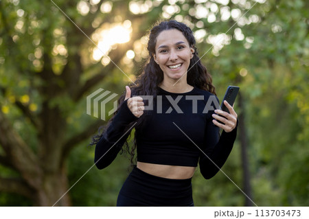 Cheerful Hispanic woman in sportswear giving a thumbs up while holding a smartphone in a lush park, conveying a message of success and connectivity. 113703473