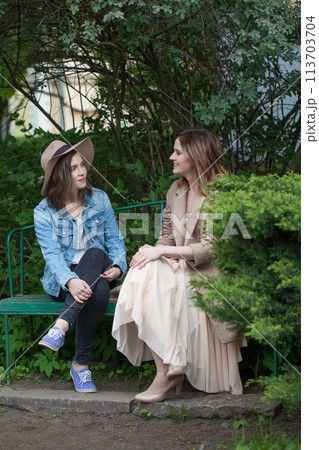 Pretty friends talking and sitting on a park bench outdoors Pretty friends talking and sitting on a park bench outdoors 113703704
