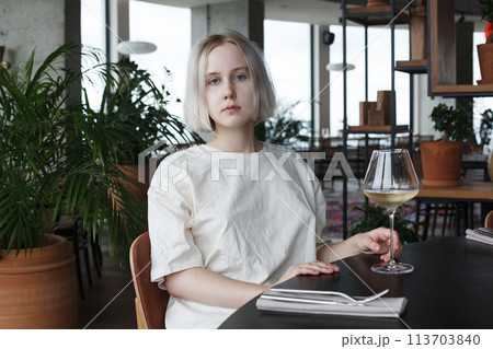 Beautiful young woman sitting at table holding in hand glass of drink at restaurant. Blonde pretty lady 113703840