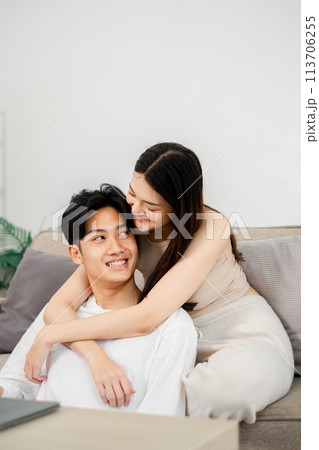 Woman in a beige dress gently embraces a smiling man in a white t-shirt from behind on a gray sofa, creating an intimate moment between them in a modern living room setting. 113706255
