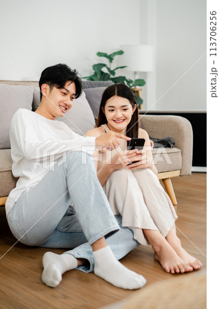 Young man in a white long-sleeve shirt and light blue jeans sits on the floor with a woman in a sleeveless dress, both enjoying content on a smartphone together. 113706256