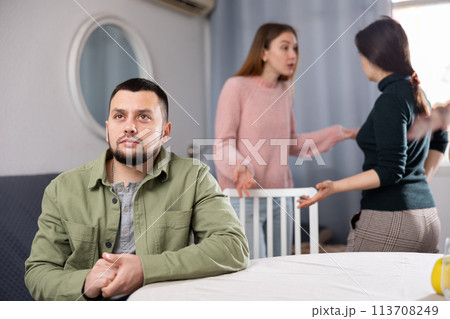 Man sitting at table while two women quarrel behind him Man sitting at table while two women quarrel behind him 113708249