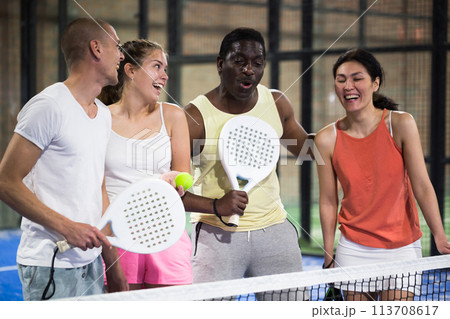 Laughing men and women with rackets and balls talking on indoor padel court 113708617