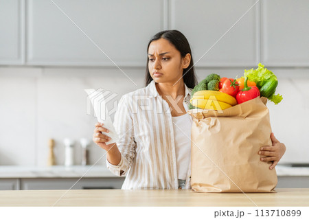 Worried Indian woman with groceries analyzing receipt 113710899