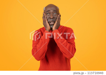 Shocked senior black man with hands on cheeks and wide eyes, wearing a red sweater 113710940