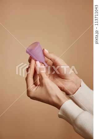 Minimal closeup of female hands holding pink menstrual cup on soft peach colored background 113711043