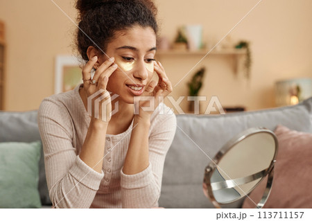Portrait of smiling young woman applying moisturizing patches and looking in mirror at home Portrait of smiling young woman applying moisturizing patches and looking in mirror at home 113711157