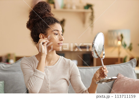 Side view portrait of young Middle eastern woman applying moisturizing face mask and looking in mirror at home 113711159