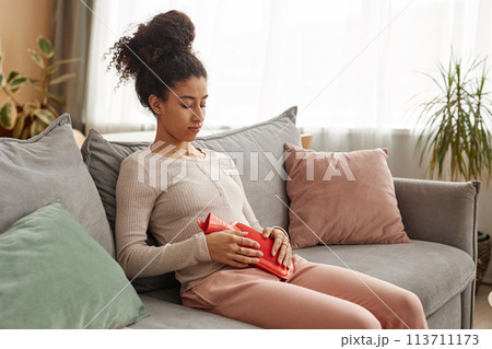 Portrait of young African American woman holding hot water bottle on belly suffering from period cramps on couch at home copy space 113711173