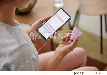 Closeup of unrecognizable girl holding menstrual cup and smartphone with period tracker app on screen copy space 113711212