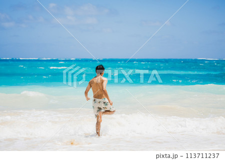 ハワイの砂浜でターコイズブルーの海に向かって走る水着姿の若い男性 113711237