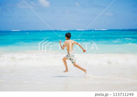 ハワイの砂浜でターコイズブルーの海に向かって走る水着姿の若い男性 113711249