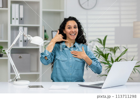 Joyful businesswoman with curly hair in a blue shirt sits at her office desk, gesturing a 'call me' sign and smiling energetically. Joyful businesswoman with curly hair in a blue shirt sits at her office desk, gesturing a 'call me' sign and smiling energetically. 113711298