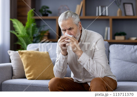 An elderly man experiencing symptoms of a cold or flu, sitting on a sofa and using a tissue in a modern, cozy living room. 113711370
