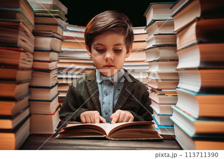 Cute little schoolboy reading book in library between stacks of books literature 113711390