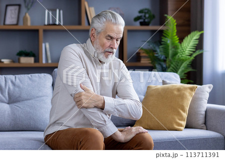 An elderly man experiencing discomfort holds his shoulder while seated on a couch, illustrating health issues in senior living. 113711391