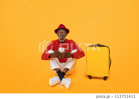 Amused senior black man in a red sweater and hat, sitting cross-legged 113711411