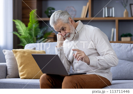 Focused elderly man with white hair and beard deeply engrossed in using a laptop while sitting on a sofa in a well-lit living room, depicting modern technology use by seniors. 113711446