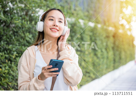 In midst of an urban city garden a happy young woman uses wireless headphones to choose and enjoy her favorite music dancing with pure joy. Her carefree spirit is a beautiful reflection of season. 113711551