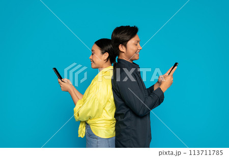 Two joyful Asian individuals, back-to-back, are smiling and engrossed in their smartphones 113711785