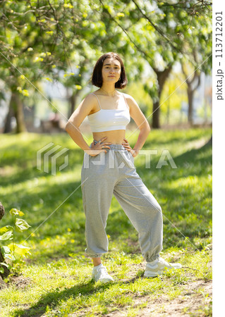 A woman is standing in a park wearing a white tank top and grey sweatpants A woman is standing in a park wearing a white tank top and grey sweatpants 113712201