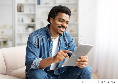 Smiling Black Man Using Digital Tablet On Couch in Bright Living Room Smiling Black Man Using Digital Tablet On Couch in Bright Living Room 113713143