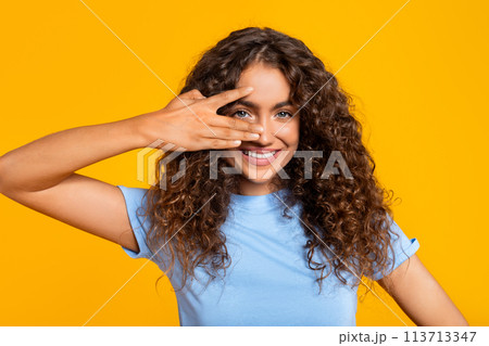 Playful woman peeking through fingers on yellow Playful woman peeking through fingers on yellow 113713347