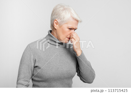 Senior woman standing with nose closed on grey background Senior woman standing with nose closed on grey background 113715425