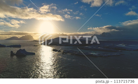 Sunset above Antarctica snow island, front aerial view. Sun reflection on ocean water in Antarctic polar summer. Beautiful sky horizon landscape. Drone forward flight 113715736