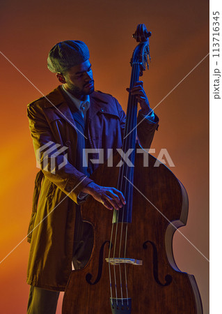 Man in retro flat cap playing double bass with vibrant blue and orange neon light against gradient background. 113716345