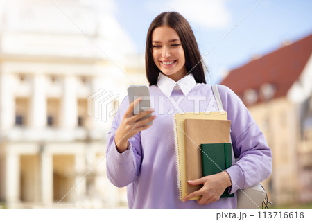 Smiling woman with books using smartphone at university campus 113716618