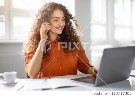 Woman conversing on phone with laptop at office 113717300