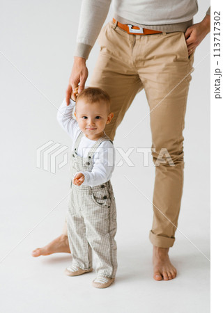 Father holds the hand of a one-year-old boy's child son teaches you how to walk. The child is wearing a white shirt and overalls Father holds the hand of a one-year-old boy's child son teaches you how to walk. The child is wearing a white shirt and overalls 113717302