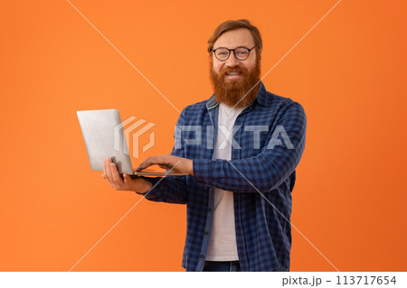 Redhaired bearded businessman works on laptop computer on orange backdrop Redhaired bearded businessman works on laptop computer on orange backdrop 113717654