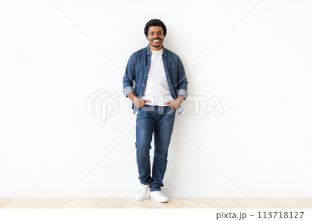 Cheerful black man standing confidently with hands in pockets against white wall Cheerful black man standing confidently with hands in pockets against white wall 113718127