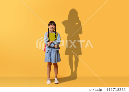 A cheerful young girl clasps a book and smiles, wearing a blue dress and backpack 113718283