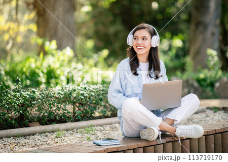 Smiling young woman sitting cross-legged on a bench in a sunlit park with headphones Smiling young woman sitting cross-legged on a bench in a sunlit park with headphones 113719170
